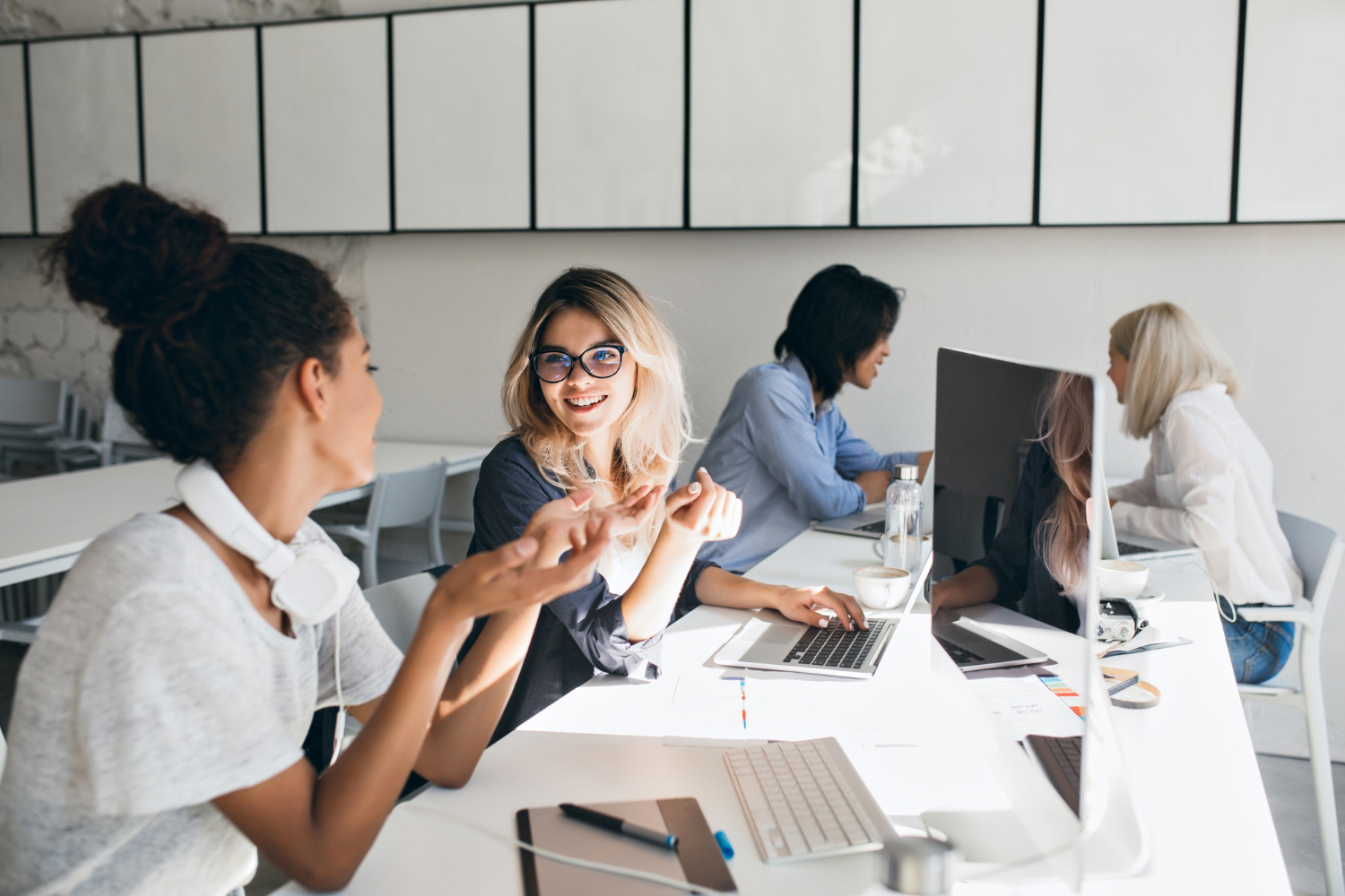 Curly-mulatto-woman-gray-t-shirt-explains-something-blonde-female-friend-indoor-portrait-international-students-with-laptops-preparing-test-together